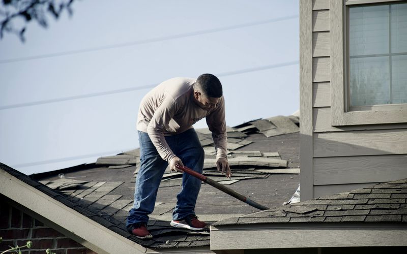 A person in a long-sleeved shirt removing shingles from a residential roof