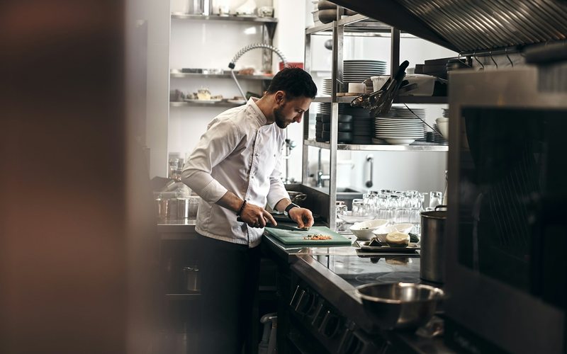 A chef in a white coat chopping vegetables on a green board in a kitchen